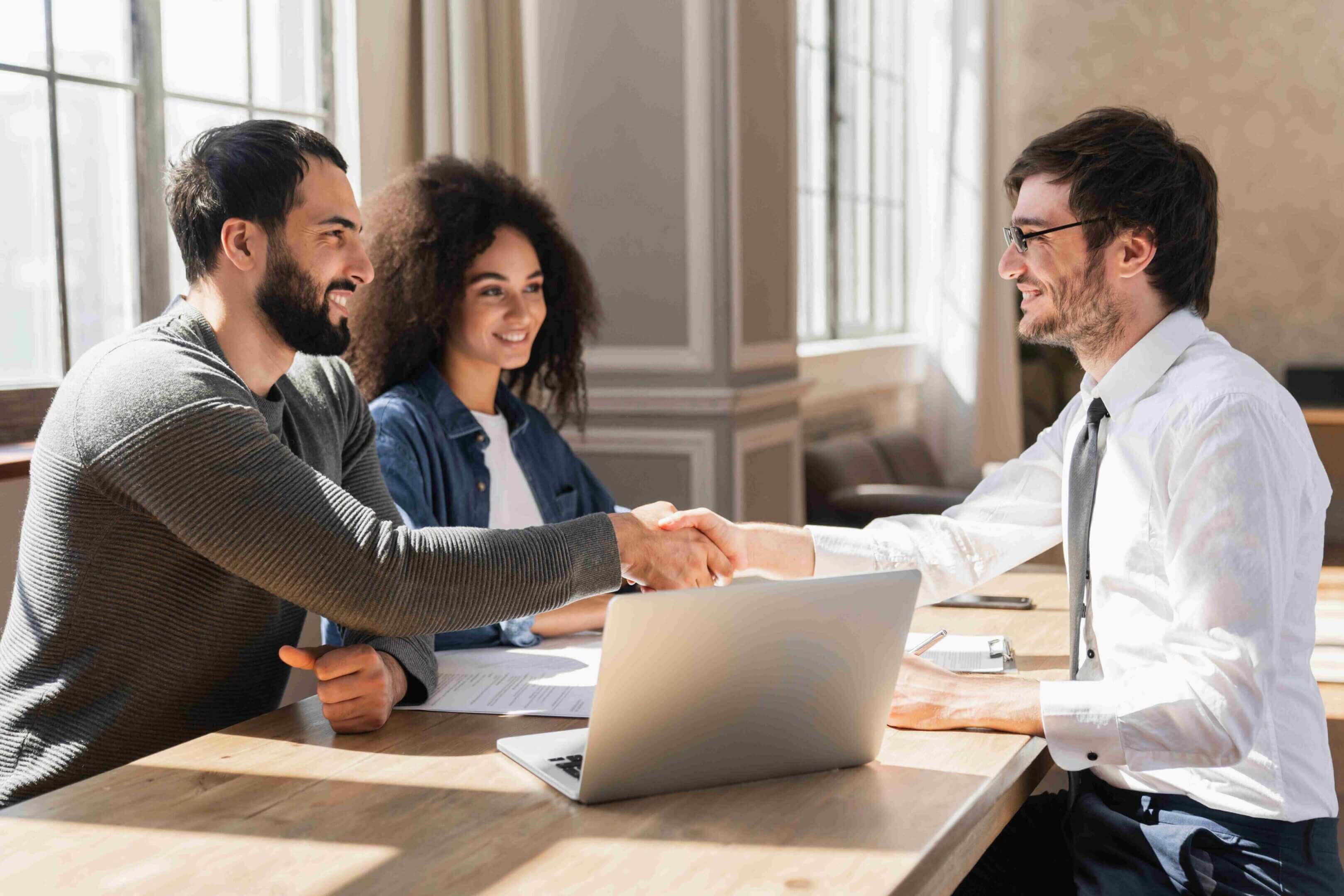 Two men shaking hands during a business meeting with a woman present.